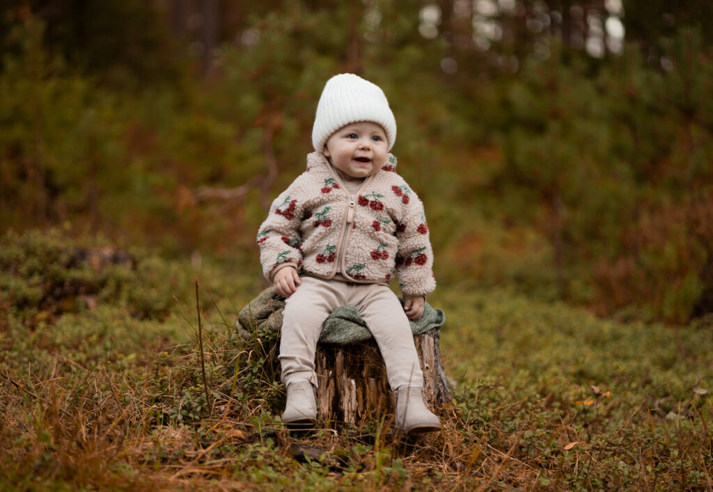 Glad bebis i mössa sitter på en stubbe i höstskogen